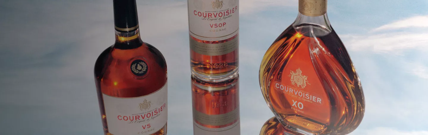 Three bottles of Courvoisier cognac set atop a reflective surface within a background of clouds.