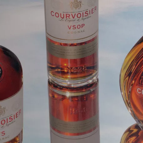 Three bottles of Courvoisier cognac set atop a reflective surface within a background of clouds.